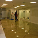 Worker and blue hose on a water-covered floor near glass partitions.