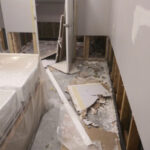 Broken wallboard and exposed lower wall studs beside covered furniture.