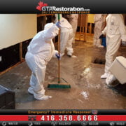 Workers in hazmat suits clean a flooded basement, using brooms to push water towards a drain, surrounded by mold removal equipment.