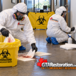 Technicians clean a small biological spill in an office corridor behind a biohazard access barrier.